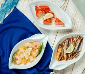 A collection of seafood dishes including shrimp with lime, seasoned anchovies, and crab meat, displayed on white plates with a nautical blue napkin.