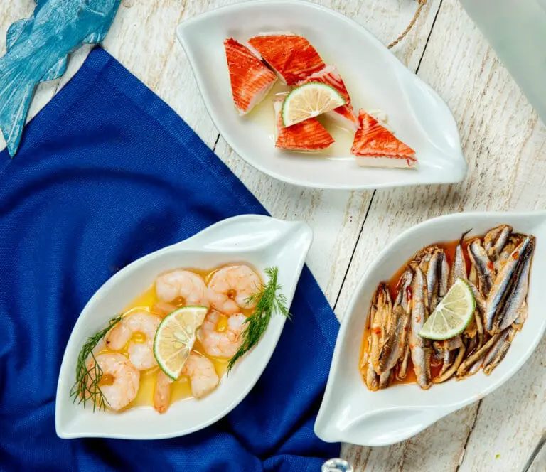 A collection of seafood dishes including shrimp with lime, seasoned anchovies, and crab meat, displayed on white plates with a nautical blue napkin.