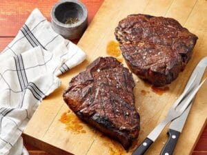 Perfectly smoked steaks on a cutting board, seasoned with coarse salt, alongside a kitchen towel and steak knives.